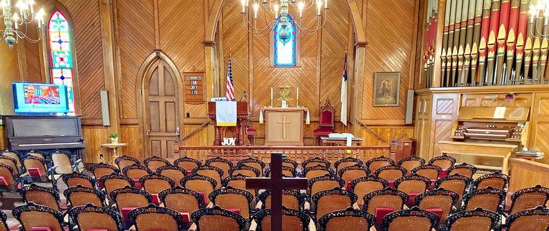 Inside the sanctuary of Rupert United Methodist Church: rows of wooden chairs facing the altar and cross, natural pine walls, stained-glass windows, and the painted pipes of the 1884 organ to the right.